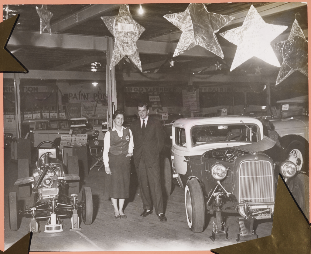 A man and a woman stand in front of two vintage cars in a warehouse with giant stars hung from the roof. 